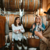 Women at an event tasting with wine bottles in front of them with barrels behind